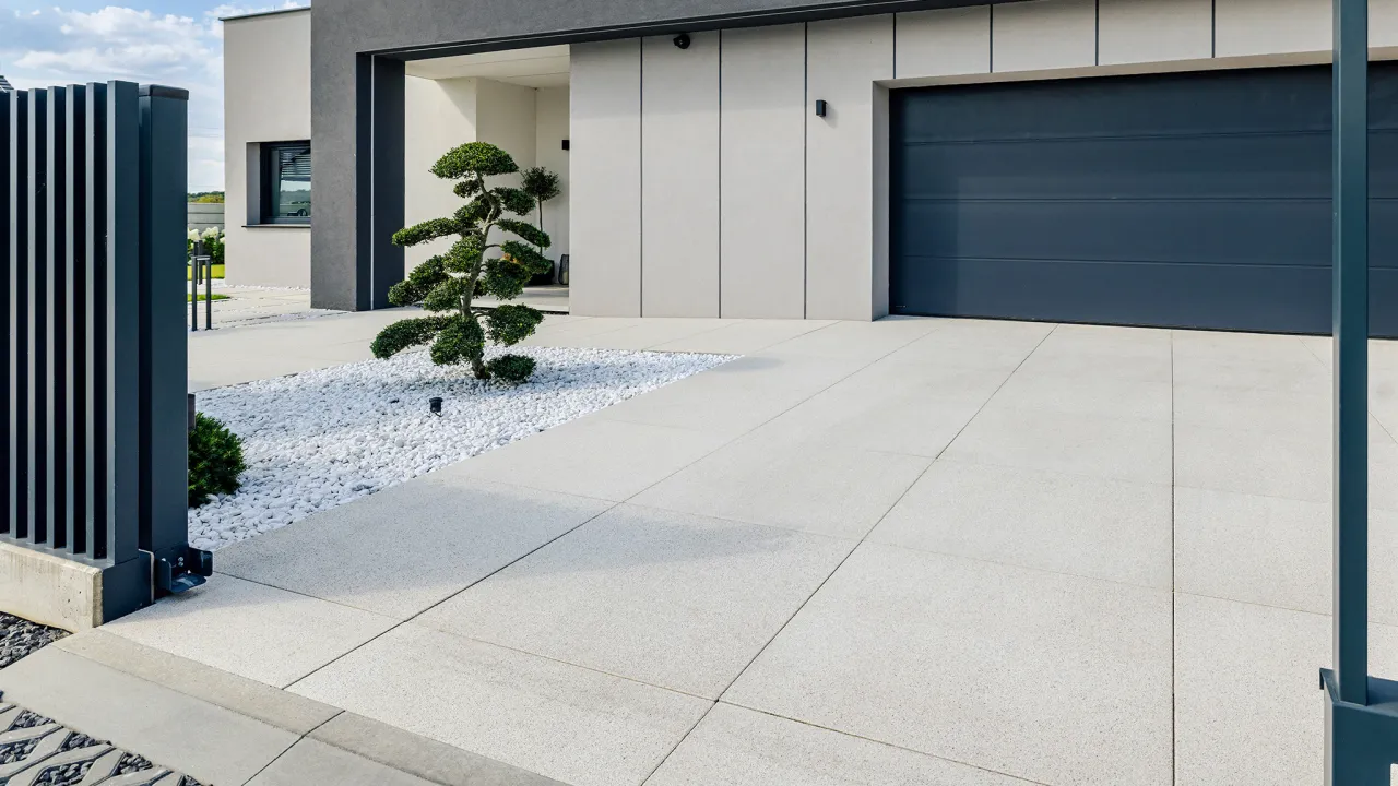 Nowoczesny podjazd przed domem z szarej kostki, ozdobiony drzewkiem bonsai na białym żwirze. Obok ciemnoszara brama garażowa.