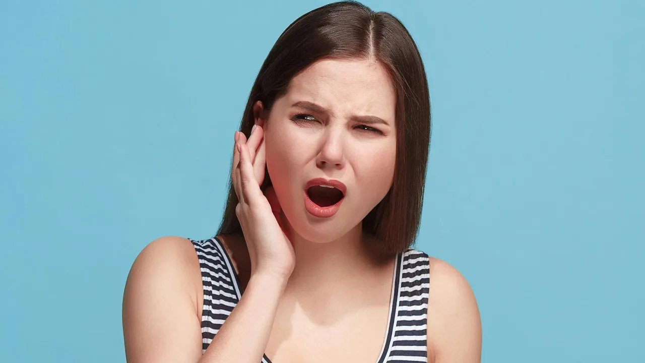Kobieta z otwartymi ustami i zmarszczonymi brwiami trzyma rękę przy uchu, zastanawiając się, dlaczego aparat słuchowy piszczy.