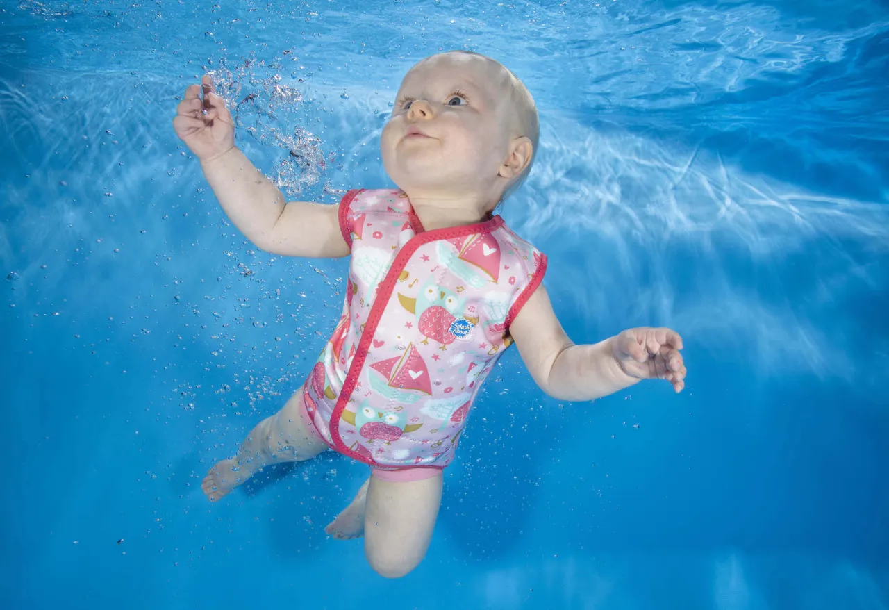 Niemowlę w r&oacute;żowym kombinezonie do pływania dla dzieci z motywem s&oacute;w i żagl&oacute;wek, unoszące się pod wodą.