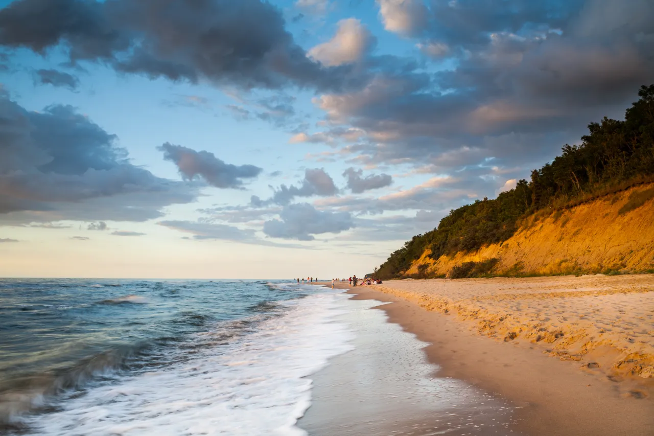 Zachód słońca nad Bałtykiem. Fale obmywają piaszczysty brzeg, a klif porośnięty drzewami mieni się złotem. To jedne z najpiękniejszych polskich plaż nad Bałtykiem.