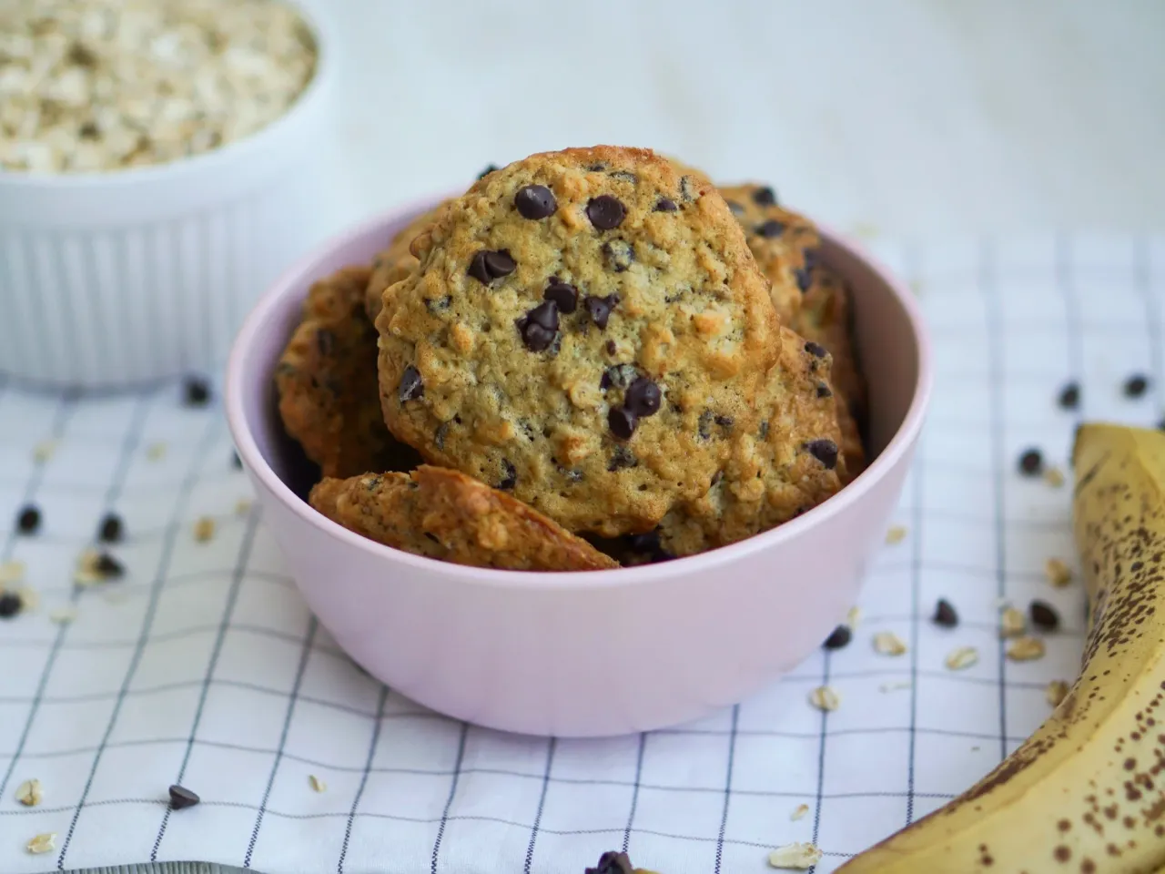 Miseczka pełna ciasteczek z kawałkami czekolady, idealnych do przepisu na zdrowe ciasteczka. Obok leży banan i płatki owsiane.