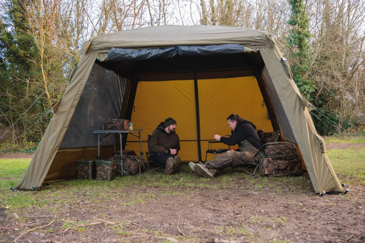 Dwóch wędkarzy siedzi w namiocie, rozmawiając. Obok nich sprzęt wędkarski, torby i stołek. To wszystko, co zabrać na ryby.