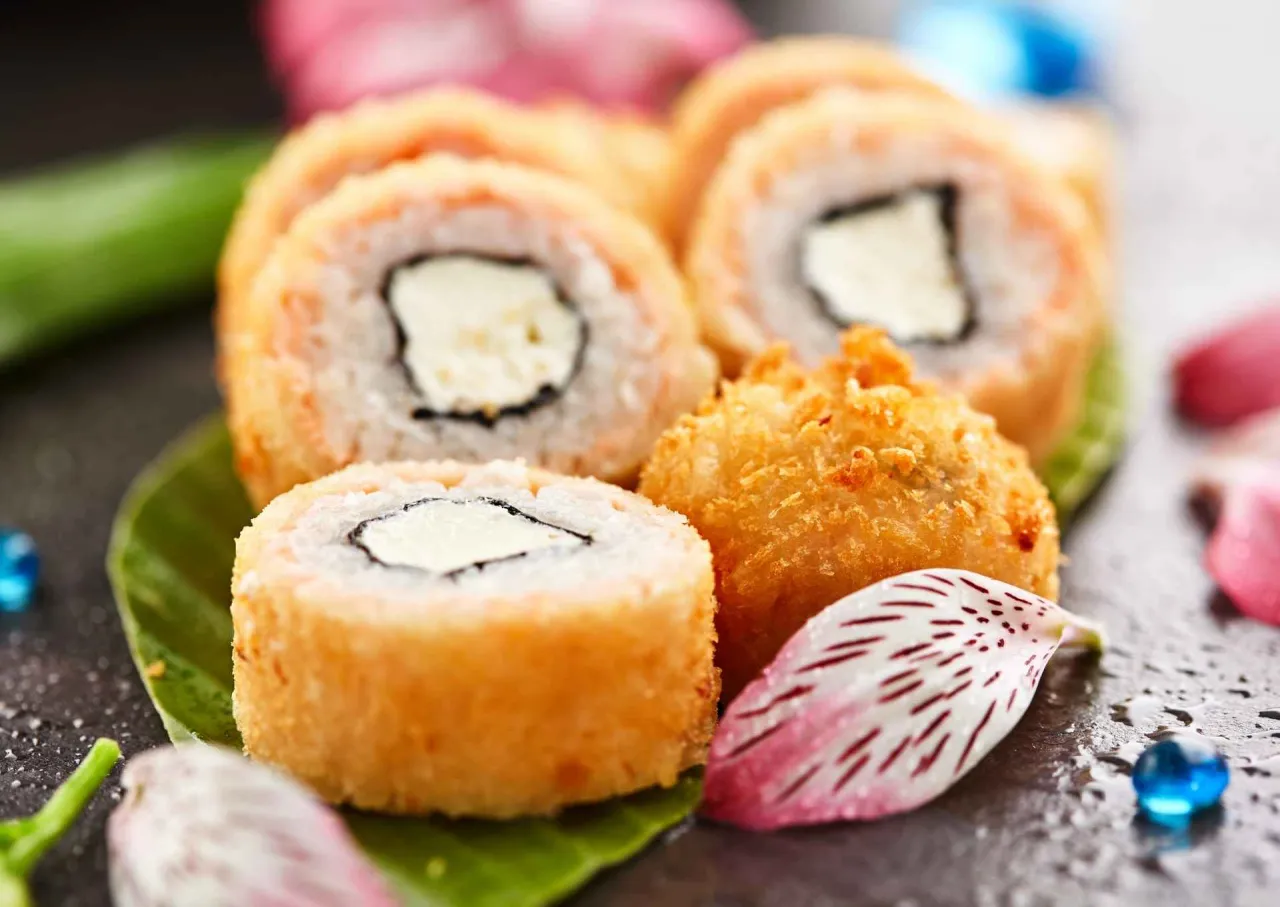 Chrupiące rolki sushi z kremowym serkiem w środku, podane na liściu, obok kwiatowych płatk&oacute;w i niebieskich koralik&oacute;w. Idealne składniki co do sushi do środka.