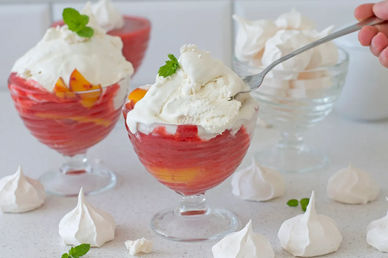 Pyszny deser Melba przepis: pucharki z musem truskawkowym, brzoskwiniami i bitą śmietaną, ozdobione miętą. Wokół bezy.