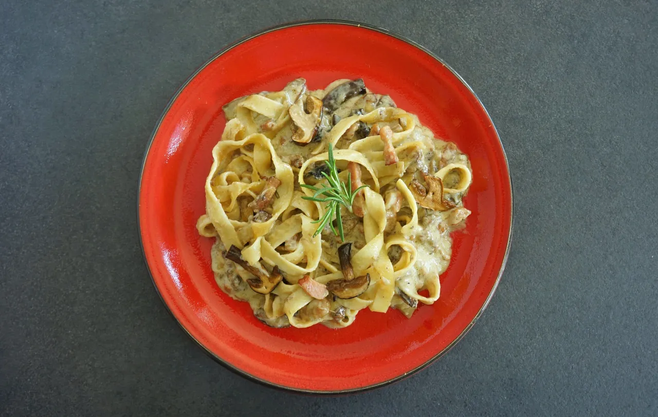 Tagliatelle z grzybami przepis. Pyszny makaron w kremowym sosie z kawałkami grzybów i boczku, udekorowany gałązką rozmarynu.