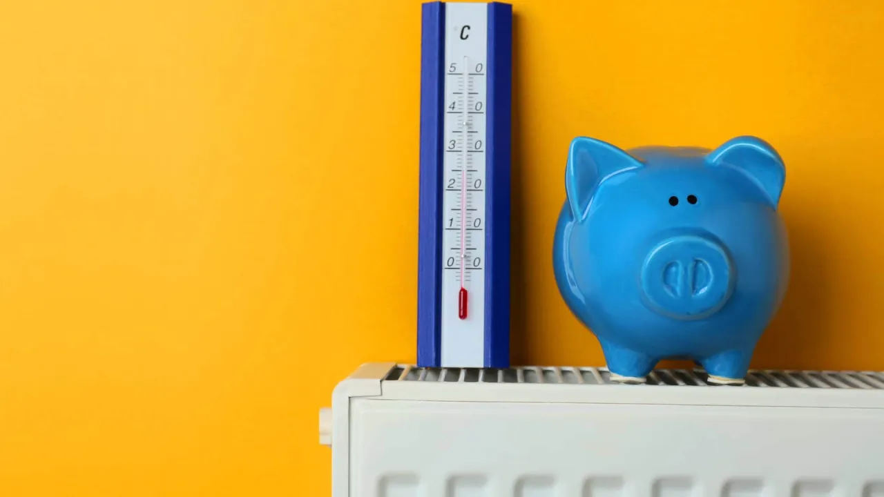 Niebieskowa świnka skarbonka i termometr na grzejniku sugerują, jaka powinna być temperatura w mieszkaniu, by oszczędzać.