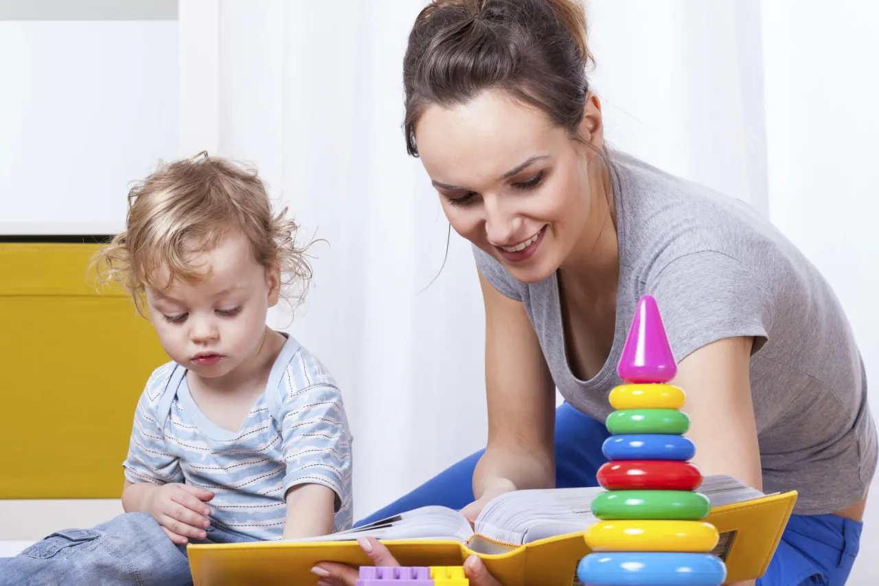 Kobieta czyta książkę dziecku, obok kolorowa wieża z kółek. Zastanawiasz się, ile kosztuje opieka nad dzieckiem?