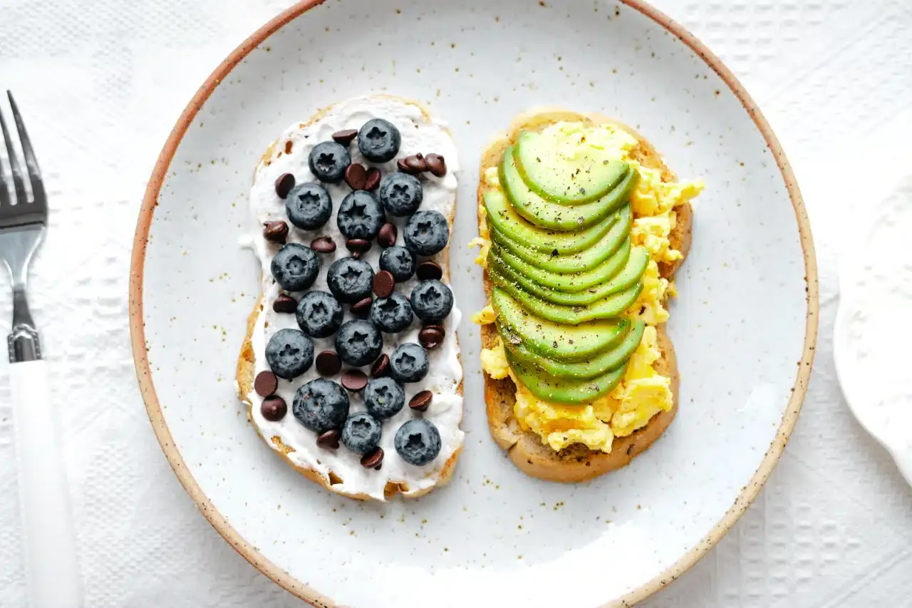 Dwa tosty na talerzu: jeden z kremem, jagodami i czekoladą, drugi z jajecznicą i awokado. Idealne propozycje, co zjeść na śniadanie wegetariańskie.