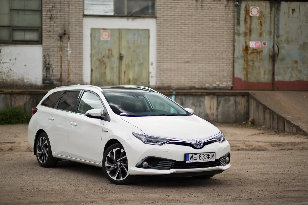 Biała Toyota Auris Hybrid kombi stoi przed zniszczonym budynkiem. Zastanawiasz się, ile pali Toyota Auris Hybrid?