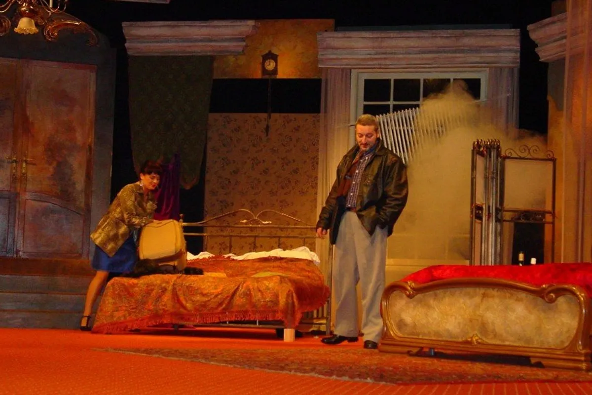 Scena teatralna z łóżkiem pełnym cudzoziemców. Kobieta z walizką przy łóżku, mężczyzna w skórzanej kurtce stoi obok.