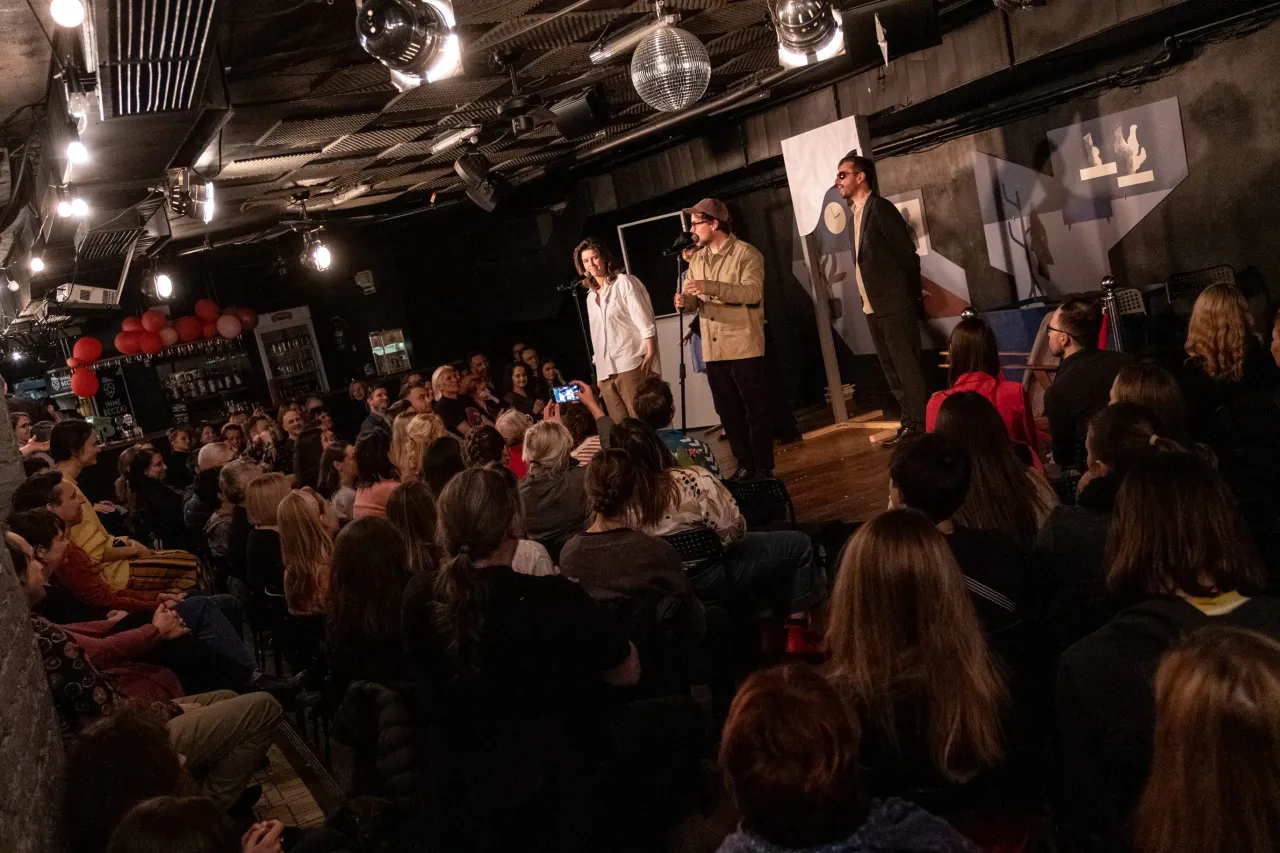 Widownia śmieje się podczas jednego z najlepszych spektakli komediowych. Na scenie trzej artyści, a przed nimi pełna sala widzów.