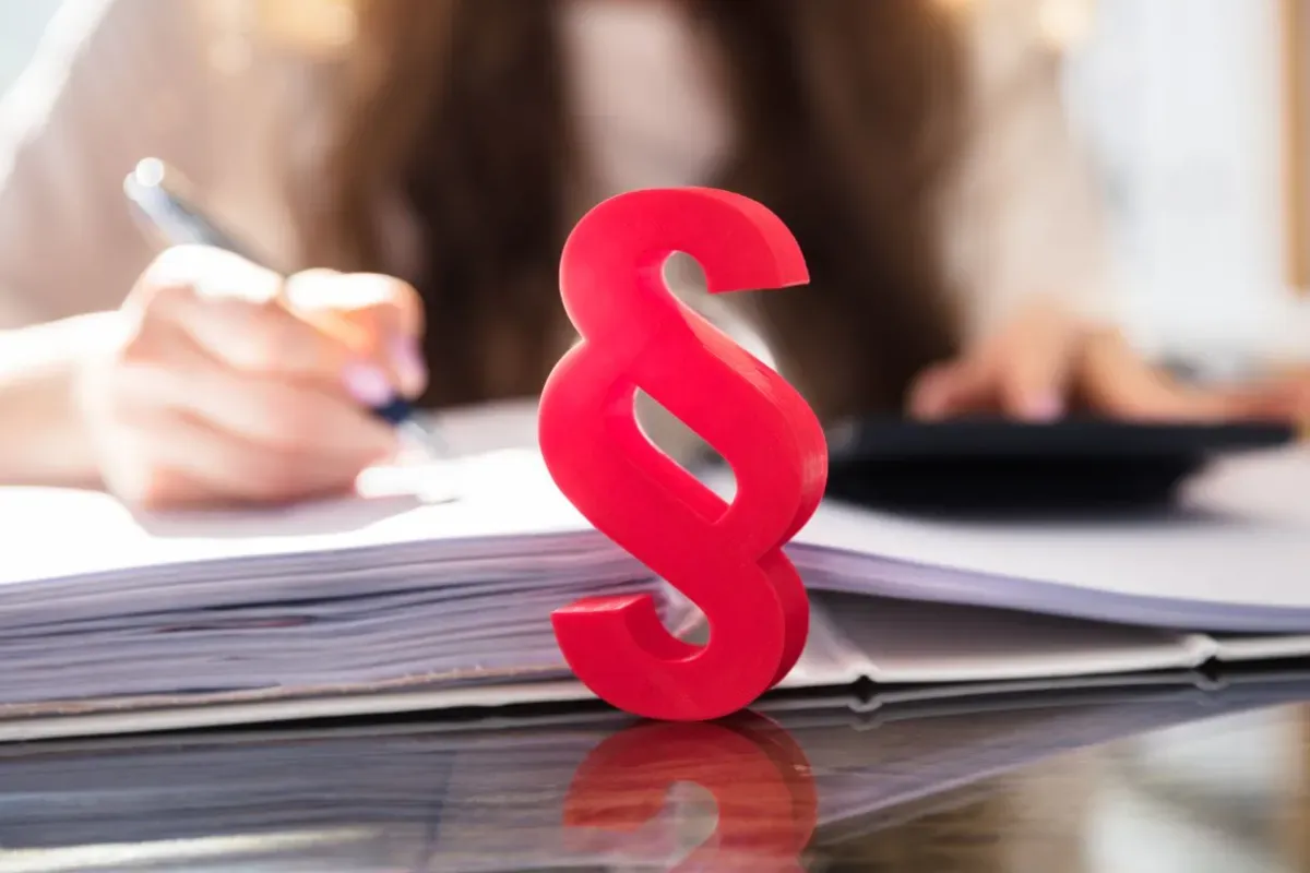 Czerwony paragraf symbolizuje prawne aspekty, gdy firma ogłasza upadłość, co z pracownikami? Tło pokazuje ręce pracujące nad dokumentami i kalkulatorem.