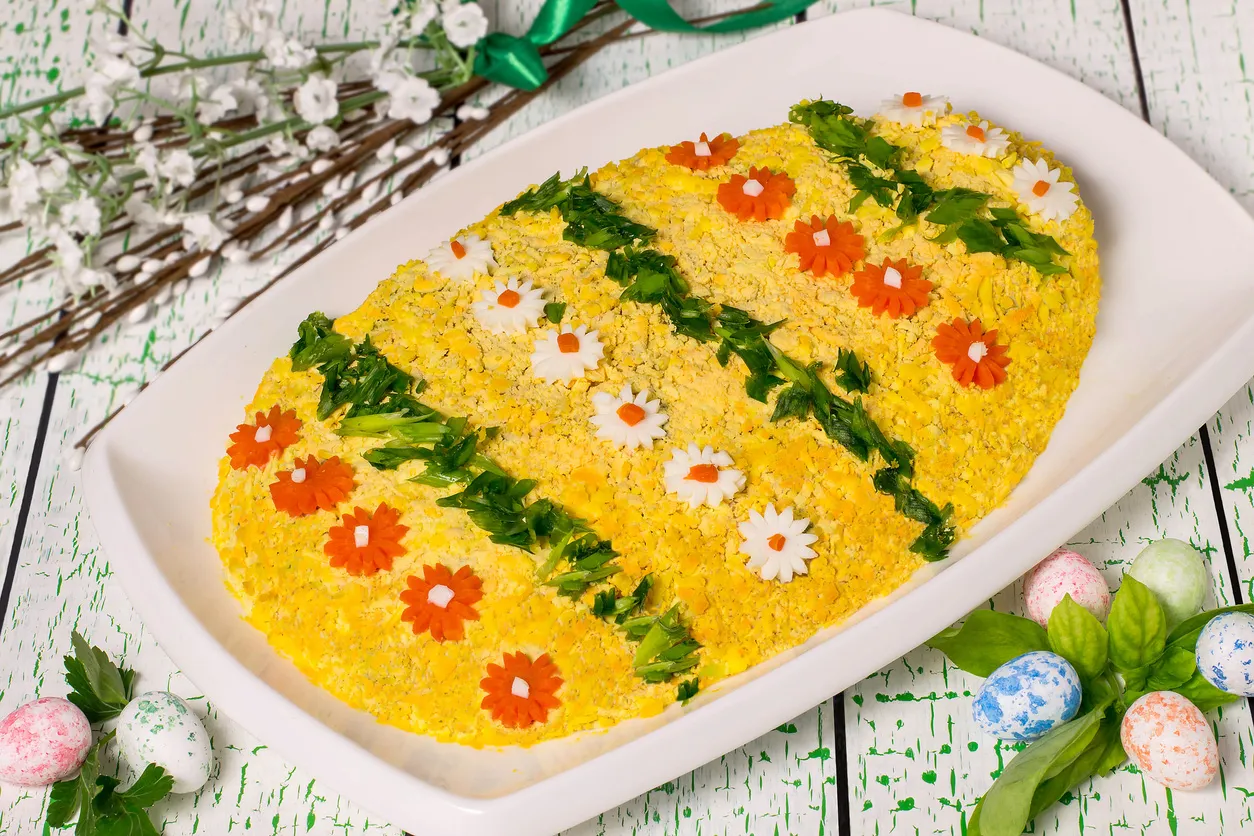 Wyborna sałatka wielkanocna w kształcie jajka, ozdobiona kwiatkami z marchewki i białka, zielonymi listkami i kolorowymi pisankami.