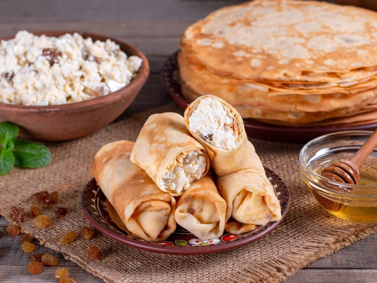 Naleśniki z twarogiem i rodzynkami – pyszny pomysł, co można zrobić z twarogu na obiad. Obok miód i stos naleśników.