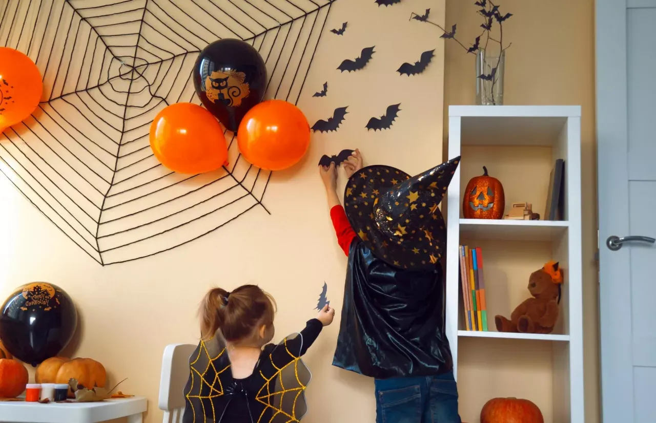 Dzieci dekorują pok&oacute;j na Halloween. Na ścianie wisi wielka pajęczyna, balony i nietoperze.