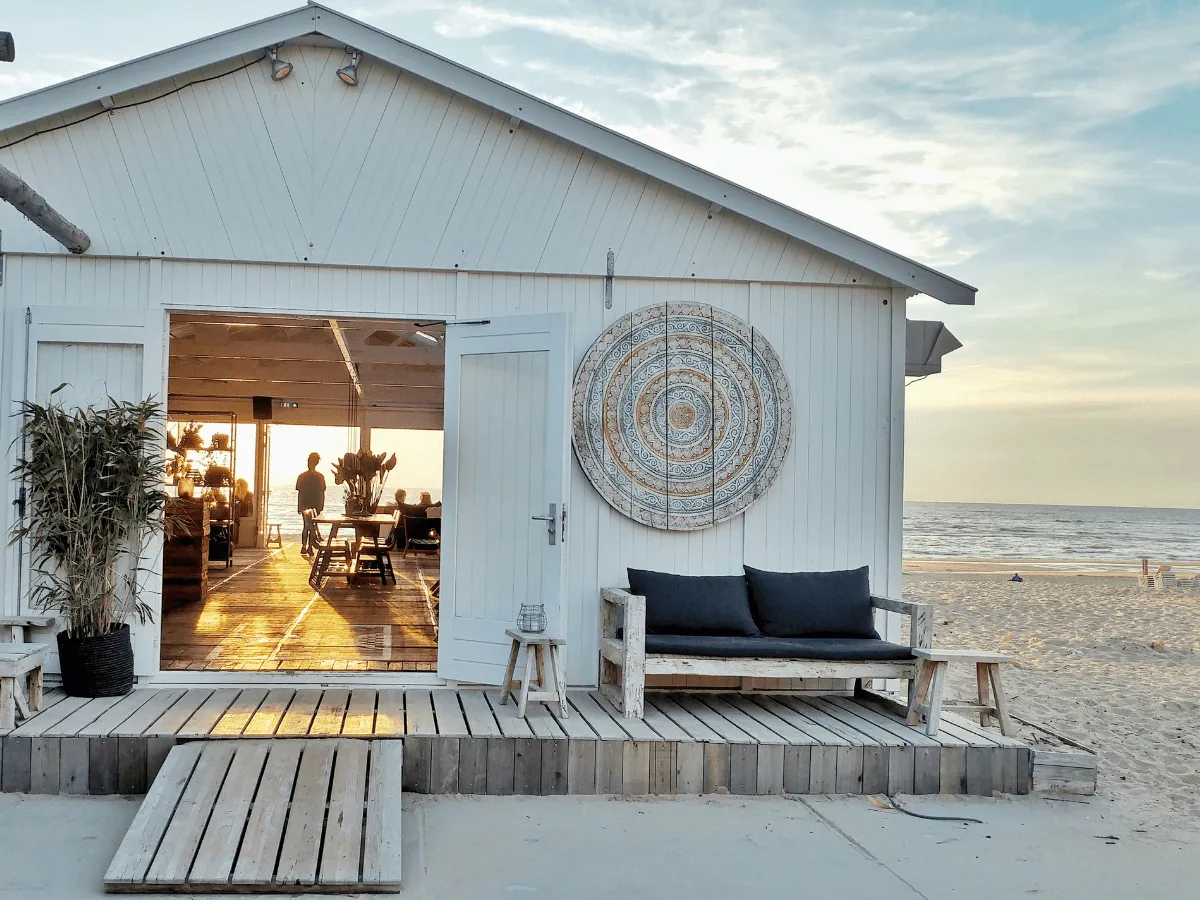 Klimatyczne noclegi nad morzem: biały domek z otwartymi drzwiami, taras z sofą i widokiem na plażę i morze.