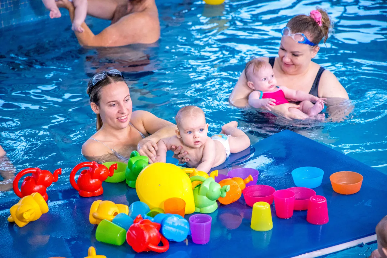 Radość z nauki pływania! Dzieci bawią się w wodzie z kolorowymi zabawkami, a rodzice pomagają im odkrywać świat basenu.