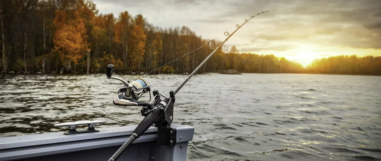 Wędka spinningowa na łodzi, gotowa do akcji. Jesienny las i zachodzące słońce tworzą idealną scenerię, by pokazać, jak łowić ryby na spinning.