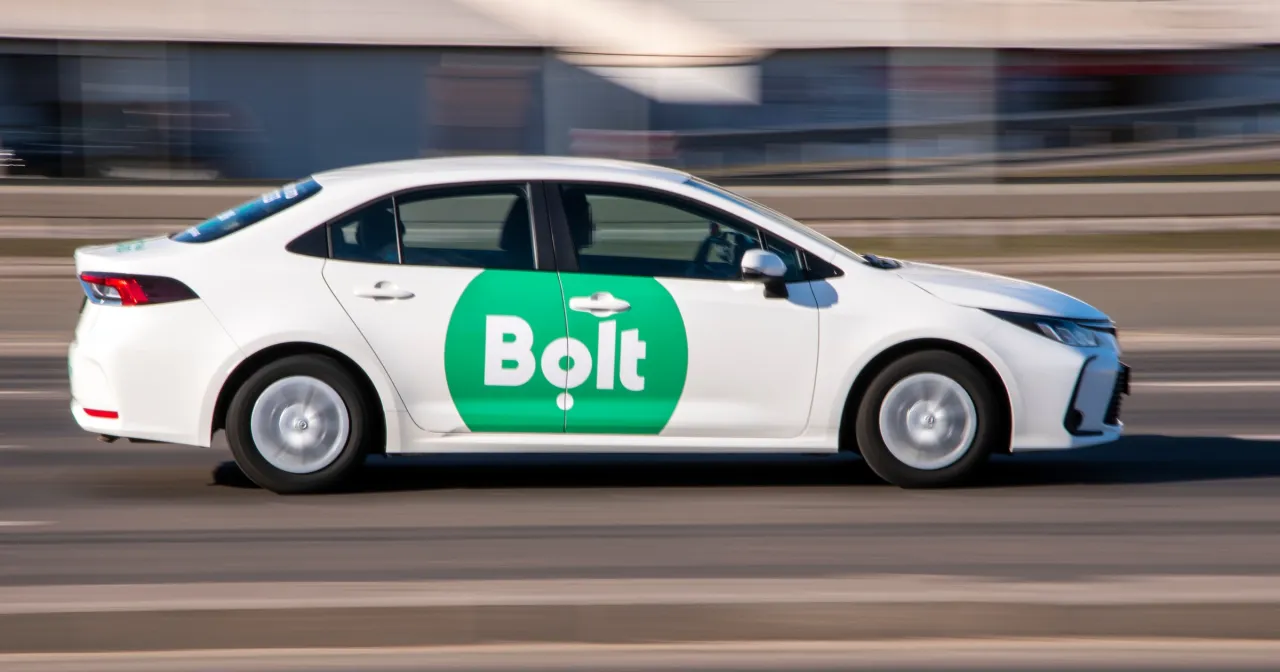 Biały samoch&oacute;d z zielonym logo Bolt na drzwiach jedzie po drodze. Chcesz wiedzieć, jak zostać kierowcą Bolta?