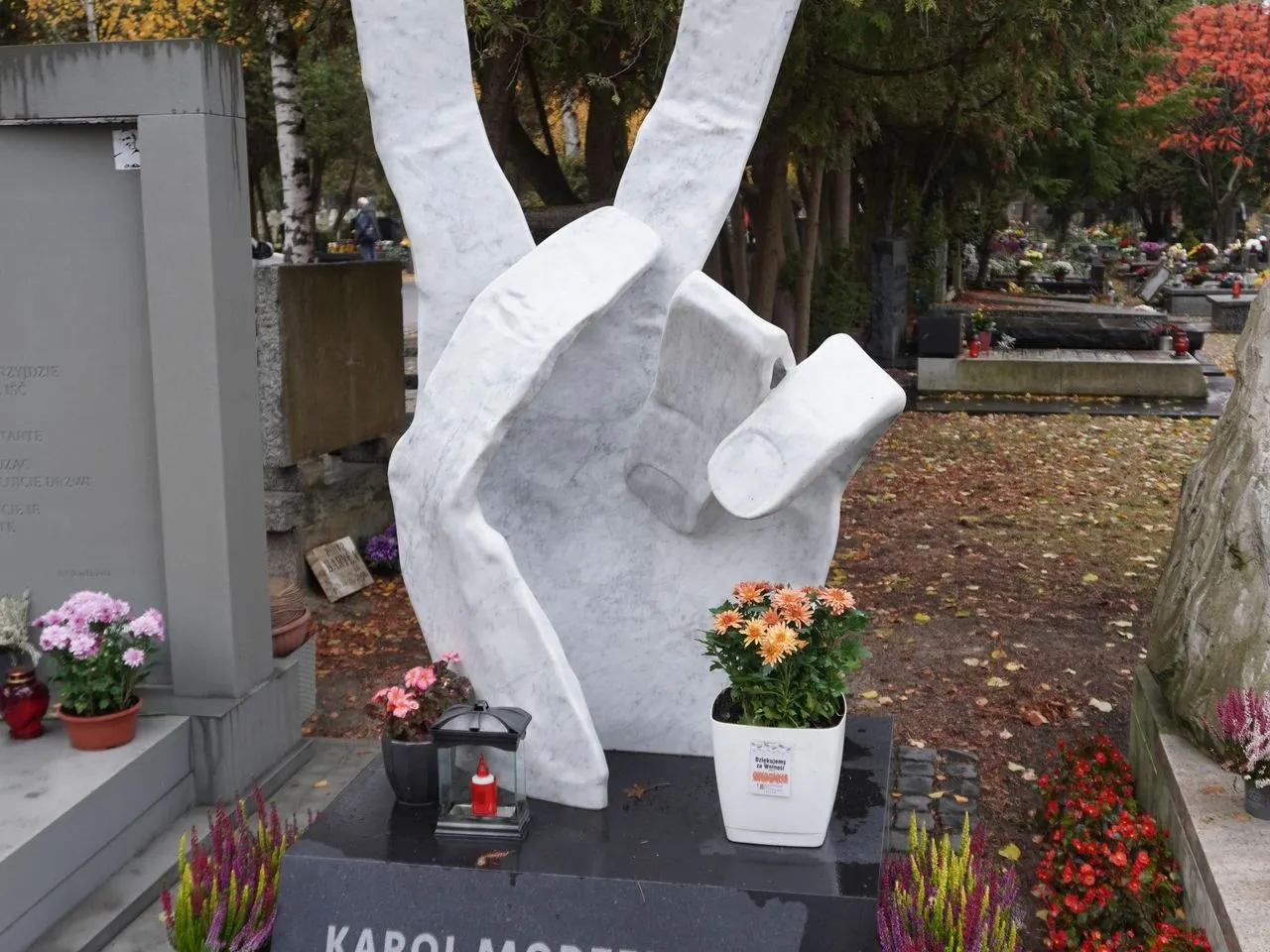 Symbol pokoju na cmentarzu w Paryżu. Biała dłoń z uniesionymi dwoma palcami, otoczona kwiatami i nagrobkami.