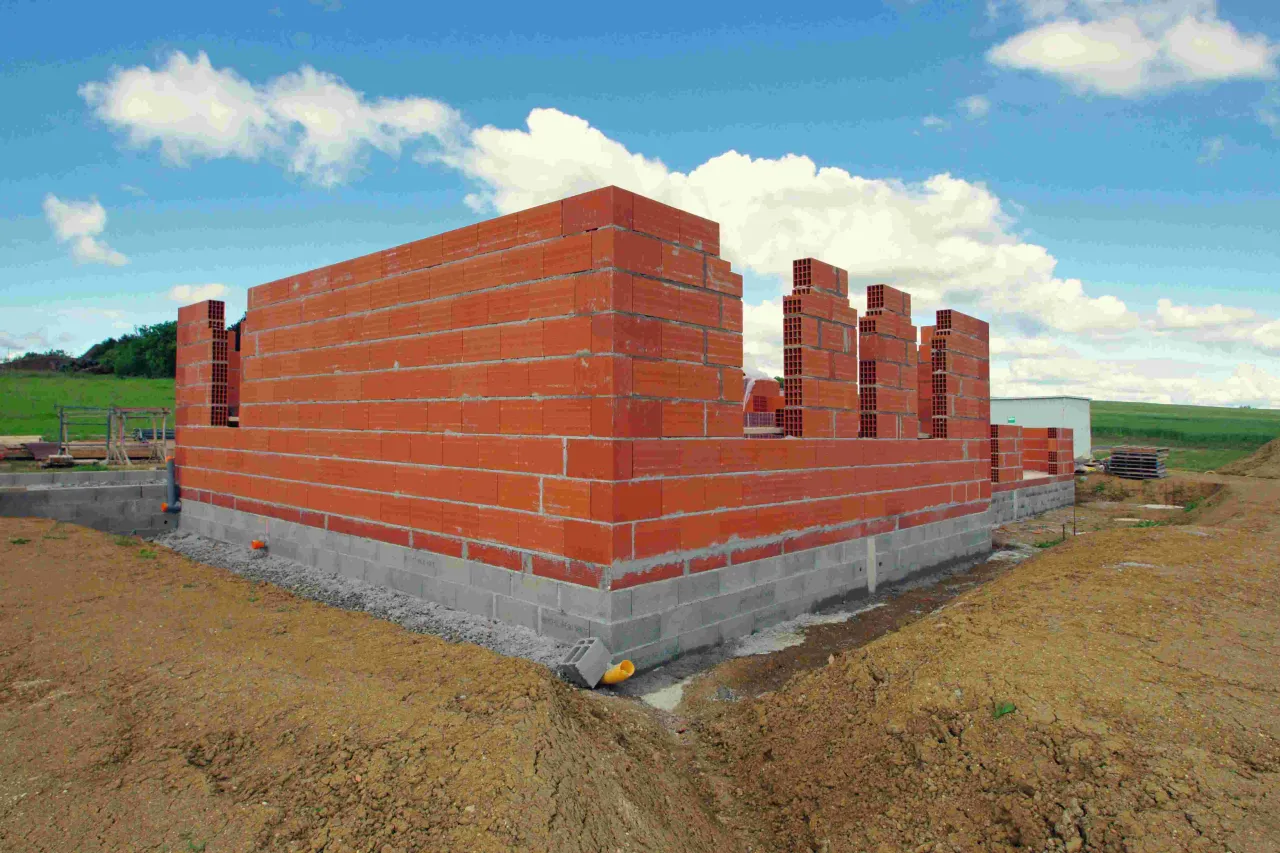 Budowa domu krok po kroku: wznoszone ściany z cegły na fundamencie z bloczków betonowych. W tle zielone pole i błękitne niebo z chmurami.