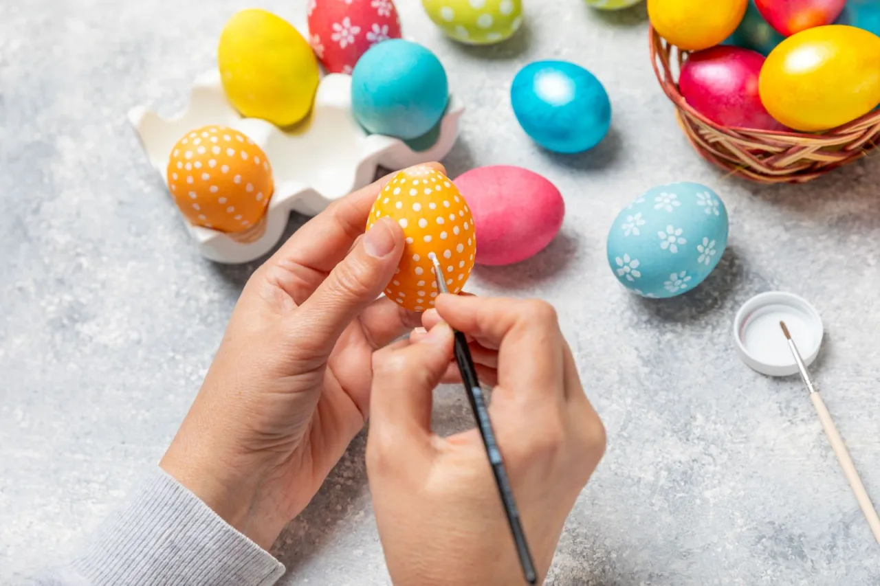 Ręce malują białe kropki na pomarańczowym jajku, tworząc piękne wzory na jajka wielkanocne. Wok&oacute;ł kolorowe pisanki.