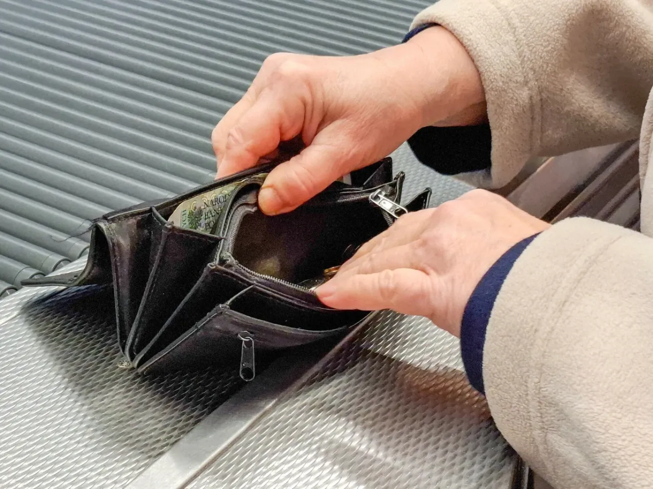 Dłonie starszej osoby przeglądają portfel, być może kiedy listonosz przynosi emeryturę. Widać kilka monet i banknot.