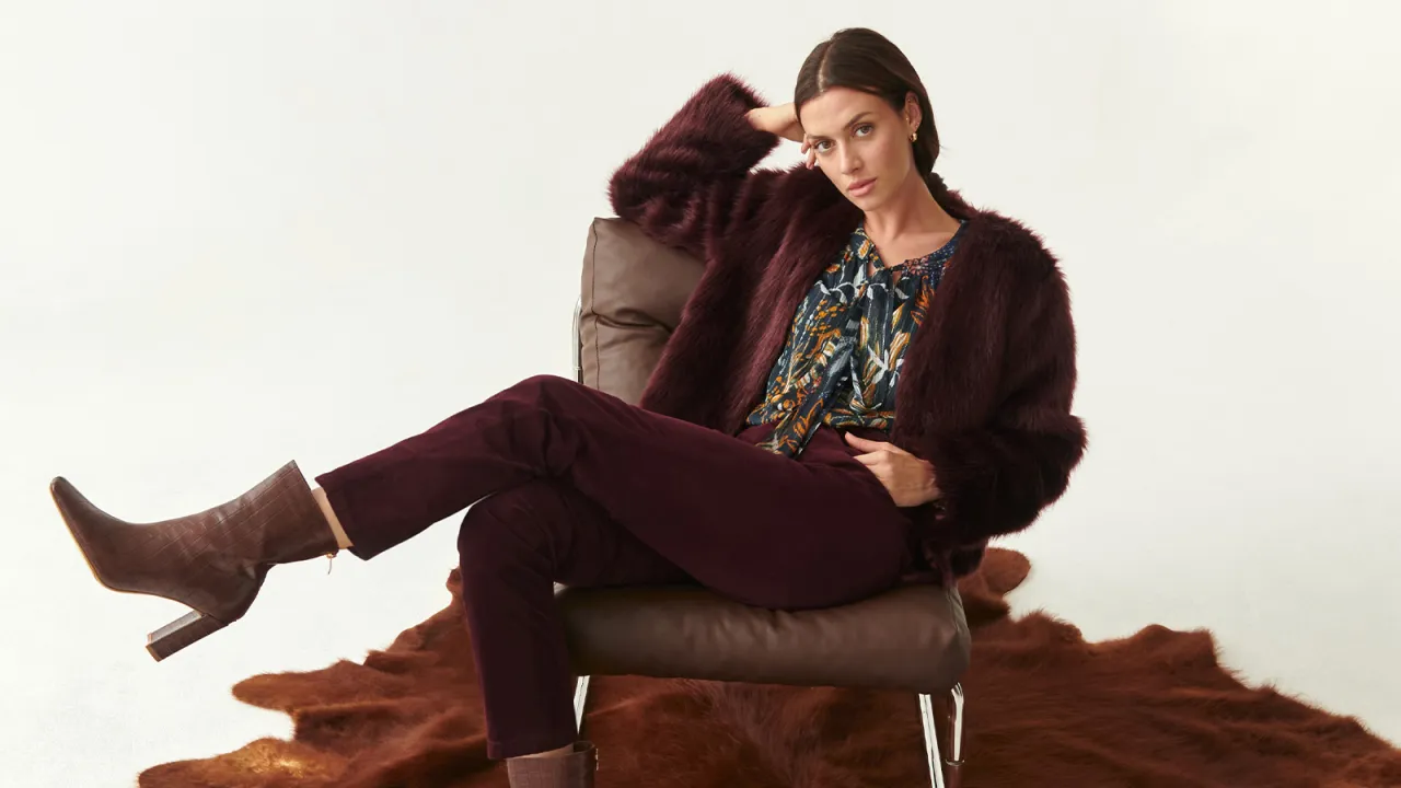 Eleganckie zimowe stylizacje damskie: bordowe sztuczne futro, wzorzysta bluzka, dopasowane spodnie i brązowe kozaki.