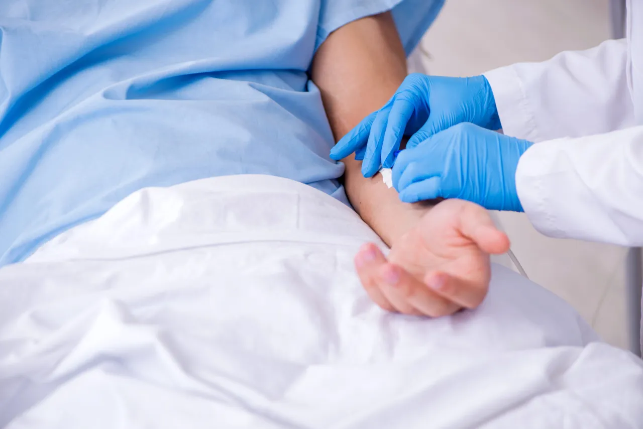 Ręce w niebieskich rękawiczkach zakładają wenflon pacjentowi. Procedura medyczna, kt&oacute;ra może nieść ryzyko czynnik&oacute;w jatrogennych.