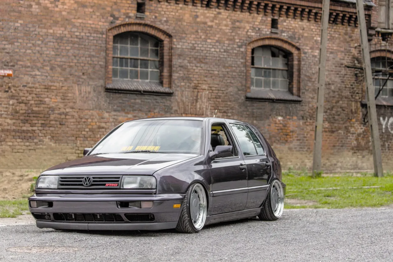 Niski, ciemny Golf R32 z felgami BBS stoi przed starą ceglaną fabryką.