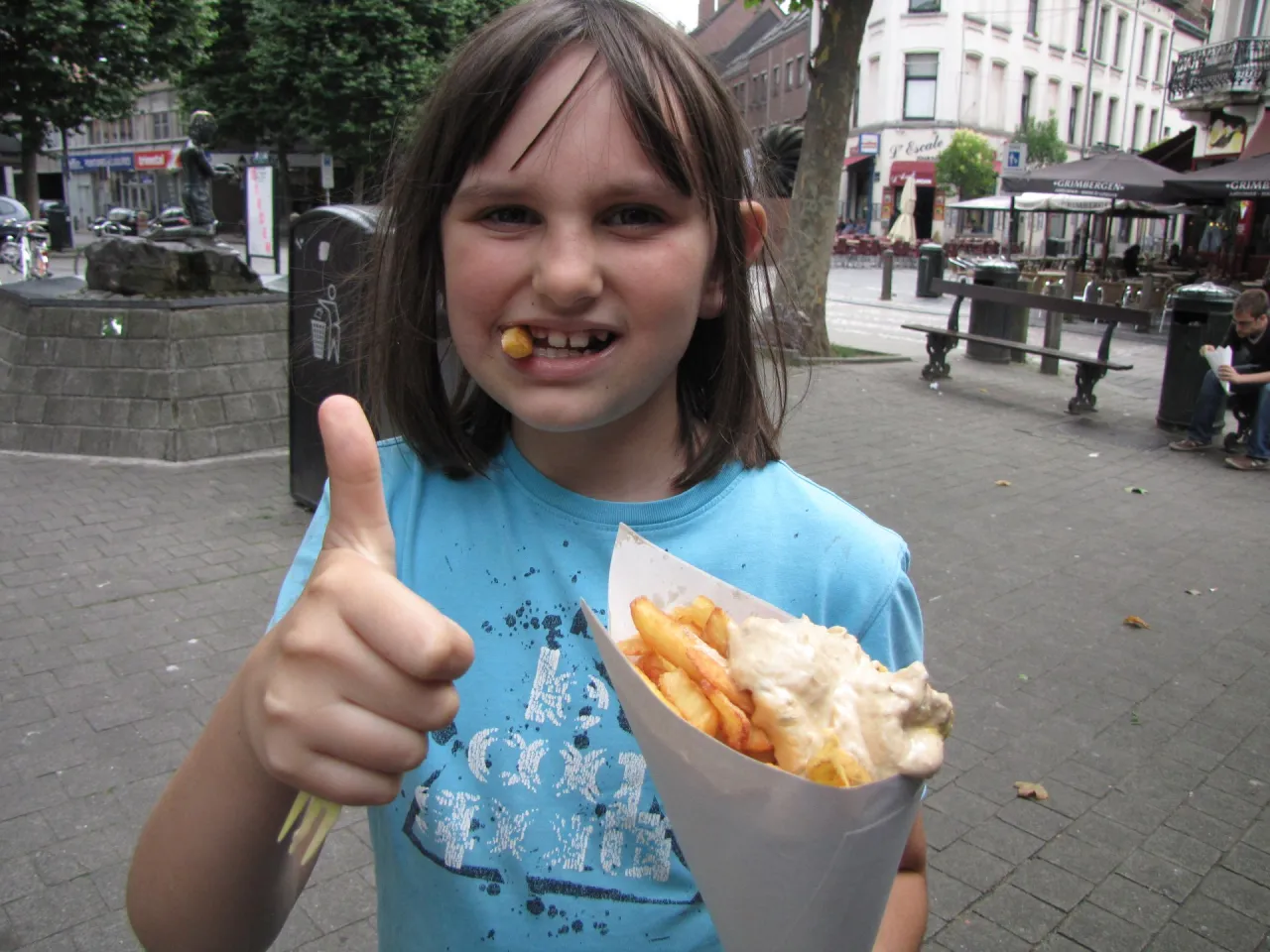 Dziewczynka z frytką w zębach pokazuje kciuk w górę, trzymając papierowy rożek z frytkami. Kto wymyślił frytki? Może ona wie!