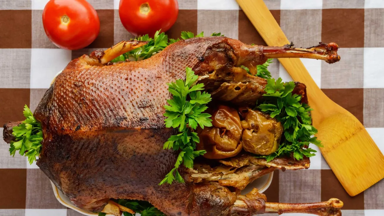 Pieczona gęś z jabłkami, ozdobiona natką pietruszki, obok pomidorów i drewnianej łopatki.