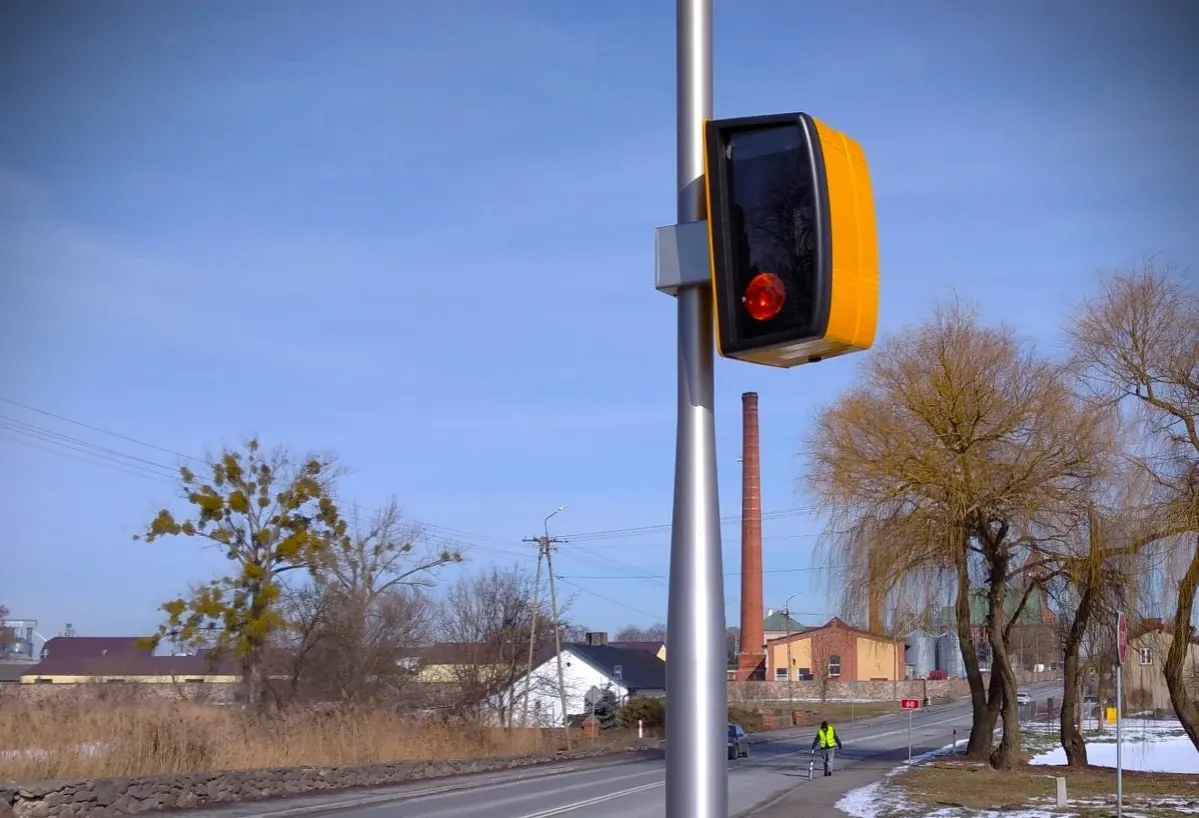 Nowe fotoradary na drodze, obok ż&oacute;łtego sygnalizatora. W tle ceglane kominy i drzewa.