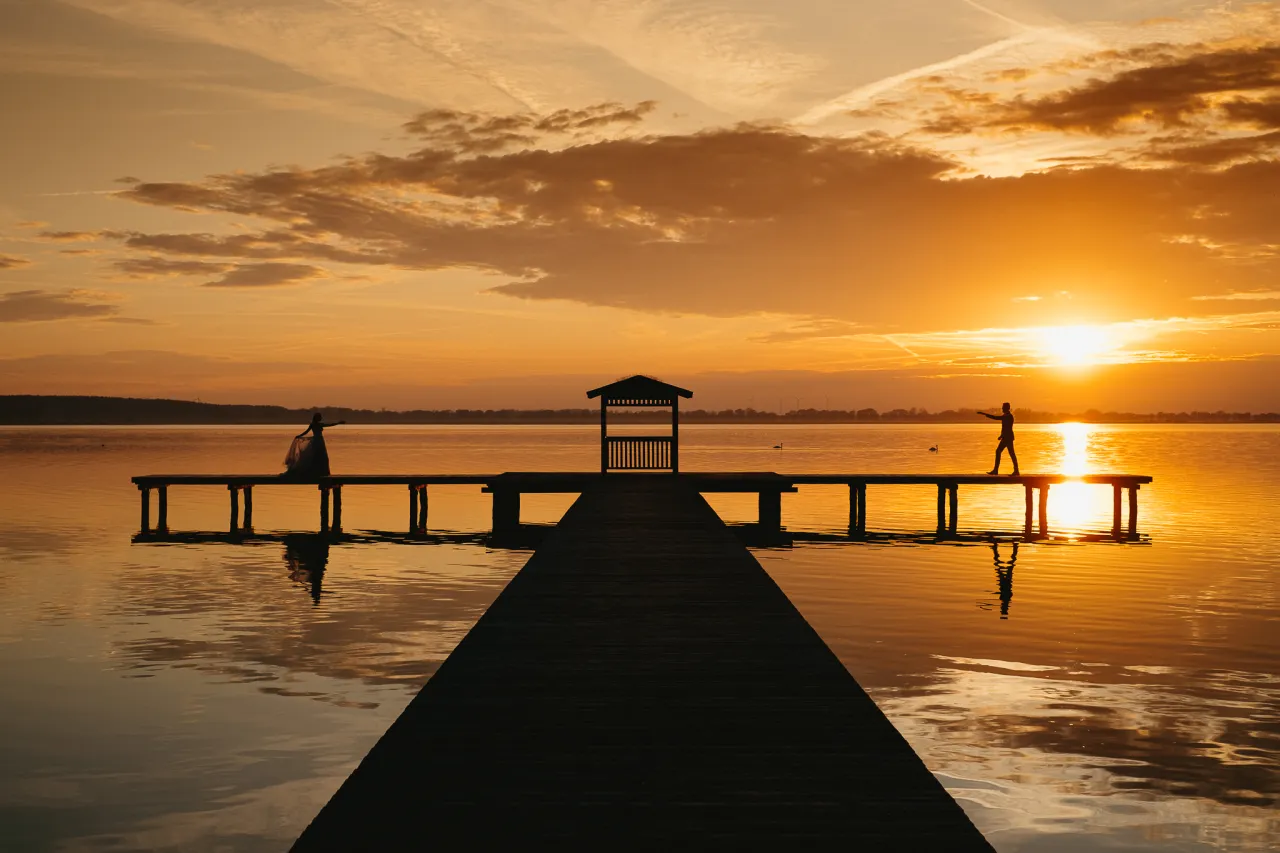 Zach&oacute;d słońca nad jeziorem, idealny moment, by nauczyć się, jak zrobić dobre zdjęcie sylwetki. Dwie postacie na pomoście.
