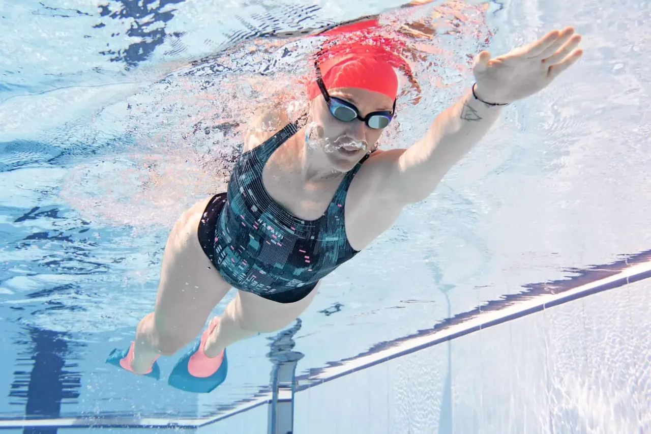 Kobieta w czerwnym czepku i okularach pływackich ćwiczy kraula, używając płetw. To świetny spos&oacute;b, jak nauczyć się pływać kraulem.