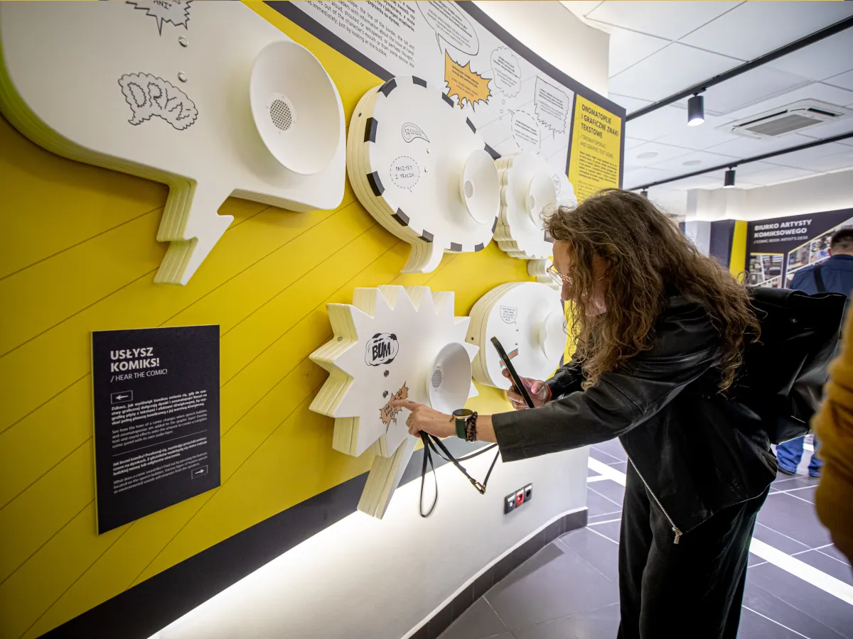 Interaktywna wystawa pokazuje, jak wygląda komiks: dymki, onomatopeje i graficzne znaki. Kobieta z telefonem bada eksponat.