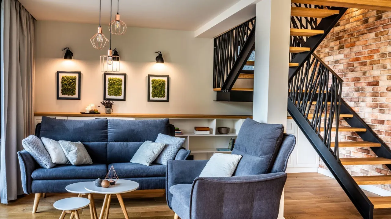 Nowoczesne wnętrze z niebieską kanapą i fotelami. Nad kanapą wiszą trzy lampy i trzy obrazy z mchem, które idealnie podkreśla ich urok. Obok schody z ceglaną ścianą.