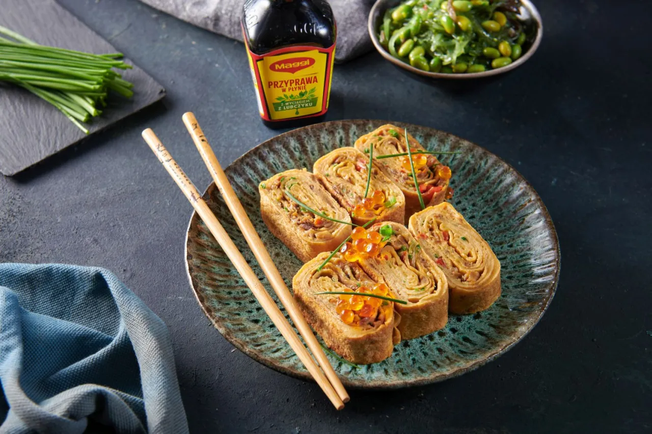 Roladki sushi w omlecie, ozdobione ikrą łososia i szczypiorkiem, prezentują się apetycznie na talerzu.