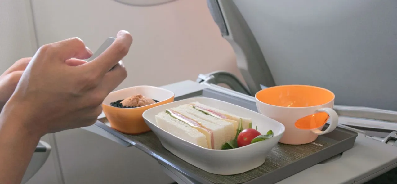 Na tacy w samolocie Ryanair leżą kanapki, deser i nap&oacute;j. Pasażer korzysta z telefonu. Czy można wziąć jedzenie do samolotu Ryanair?