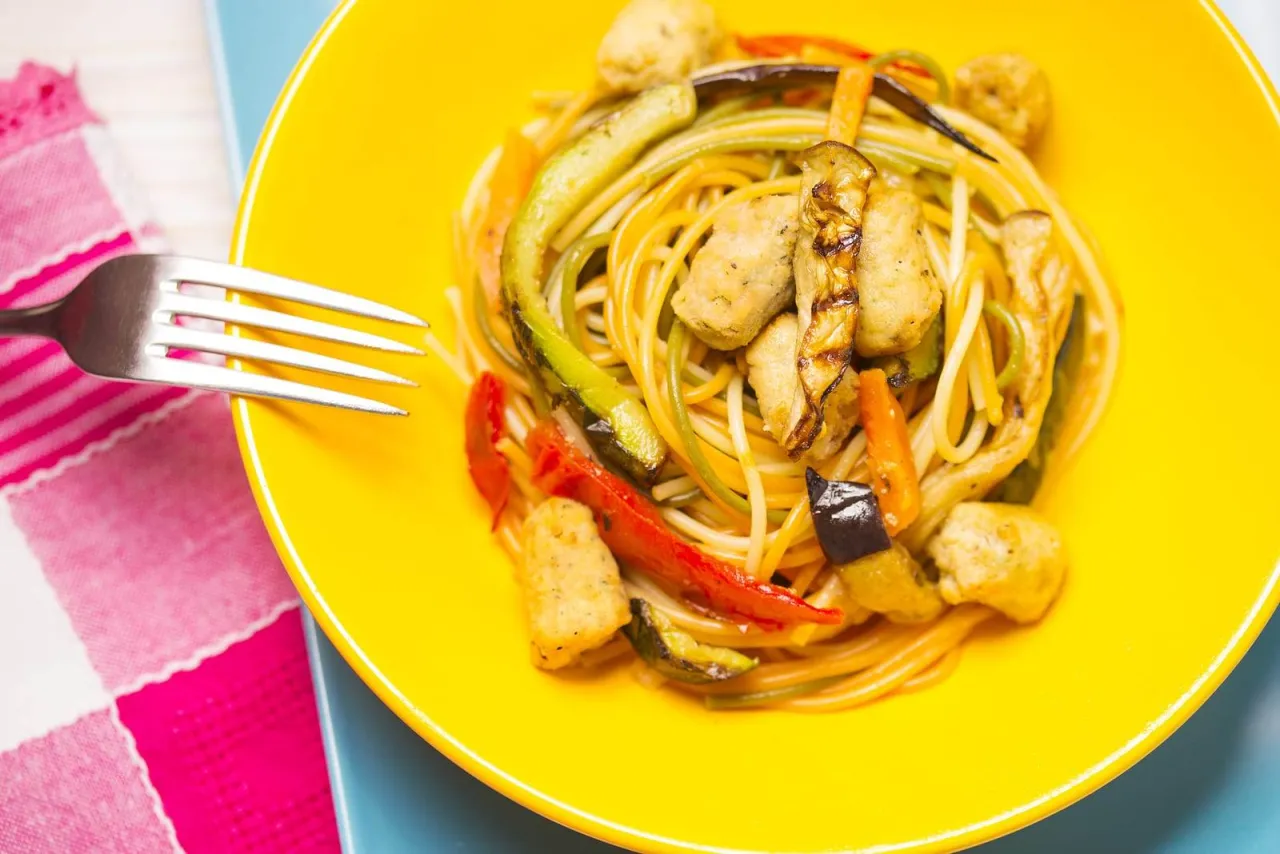 Kolorowy makaron z warzywami i kurczakiem na żółtym talerzu. Zastanawiasz się, ile kalorii ma makaron sojowy?