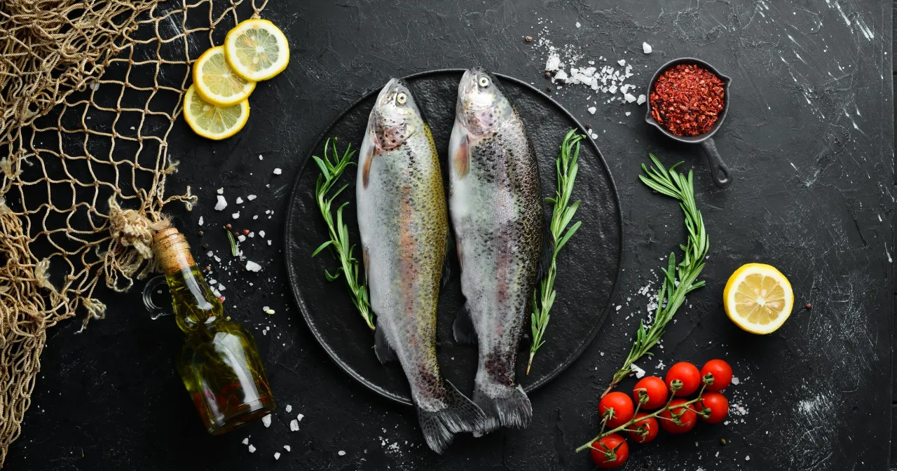 Dwa świeże pstrągi na talerzu, otoczone cytryną, rozmarynem i pomidorkami. Pstrąg wartości odżywcze są wysokie, to zdrowy wybór.