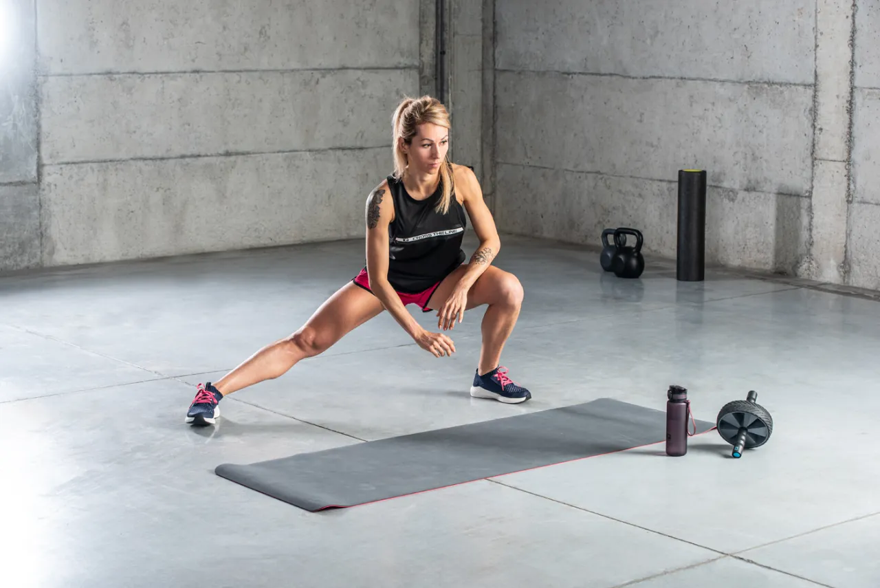 Kobieta rozciąga się po treningu, szukając sposob&oacute;w na co na zakwasy po treningu. Na macie leży butelka i przyrząd do ćwiczeń.