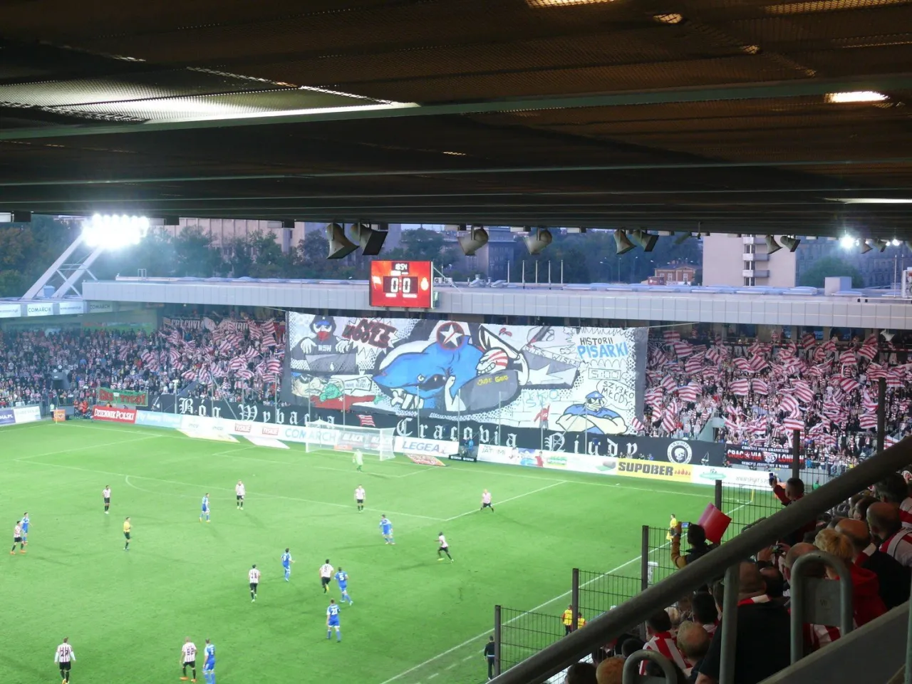 Widok stadionu podczas meczu piłki nożnej, gdzie kibice tworzą oprawę. To pokazuje, co to są derby w piłce nożnej &ndash; emocje i rywalizacja.