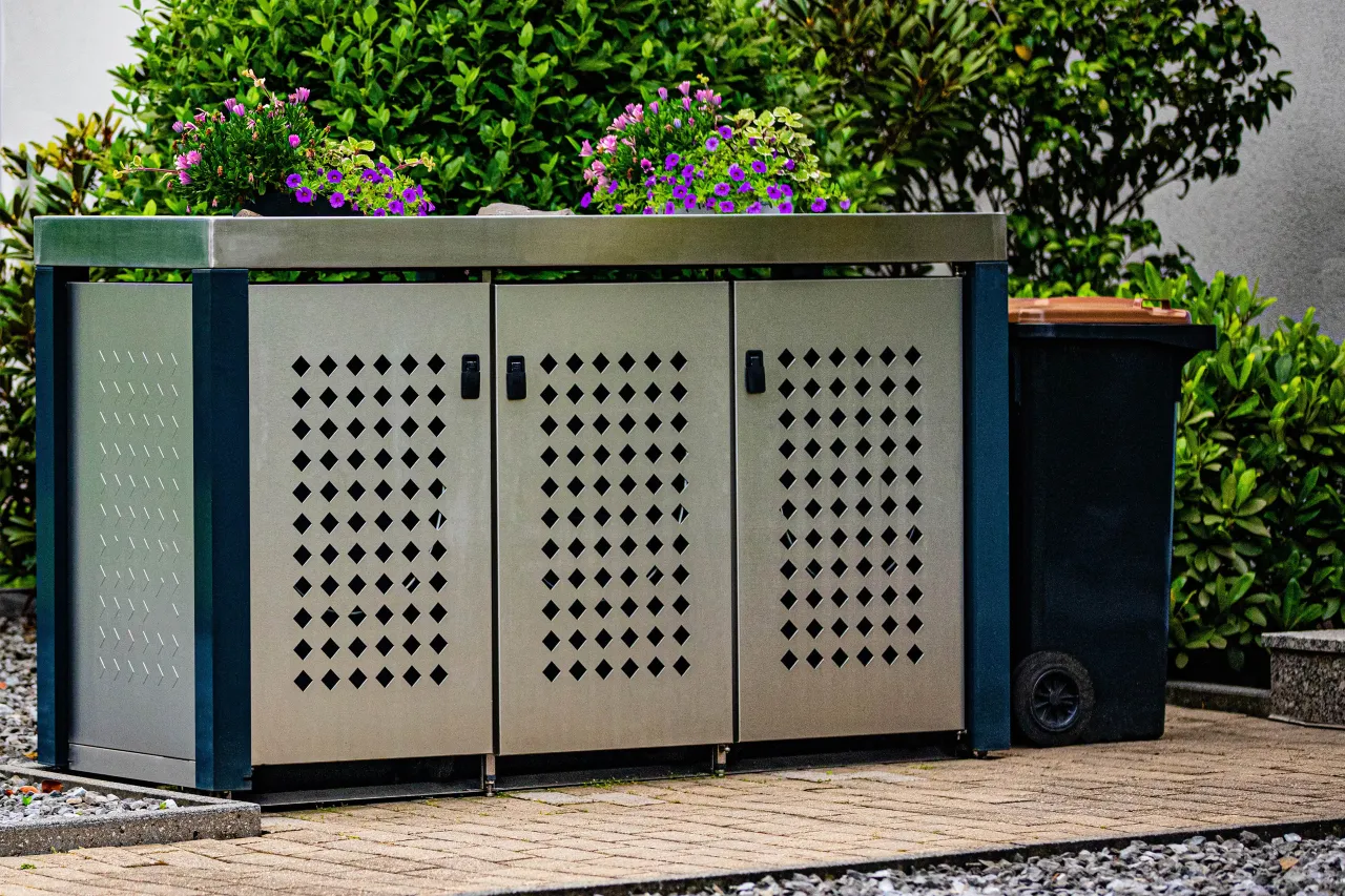 Nowoczesna zabudowa na kosze w ogrodzie, z kwiatami na g&oacute;rze, to świetny spos&oacute;b, jak ukryć kosz na śmieci.