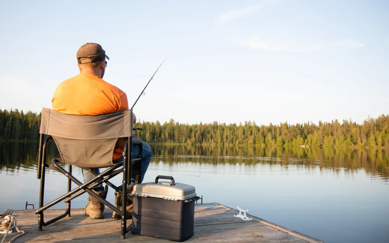 Wędkarz na pomoście, zastanawia się, ile kosztuje karta wędkarska na całą Polskę. Spokojna tafla jeziora i las w tle.