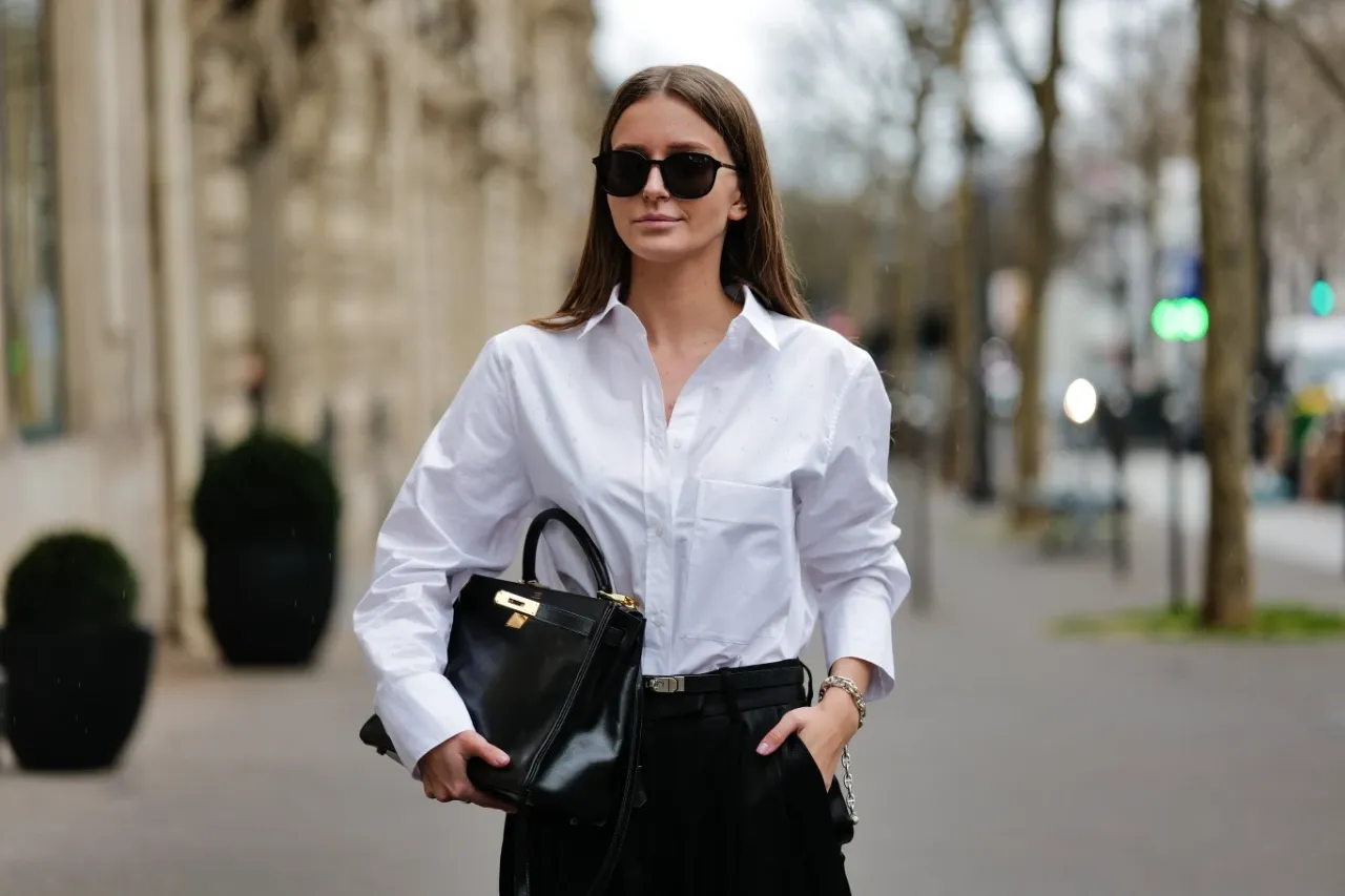 Elegancka kobieta w białej koszuli i czarnych spodniach, trzymająca czarną torebkę.