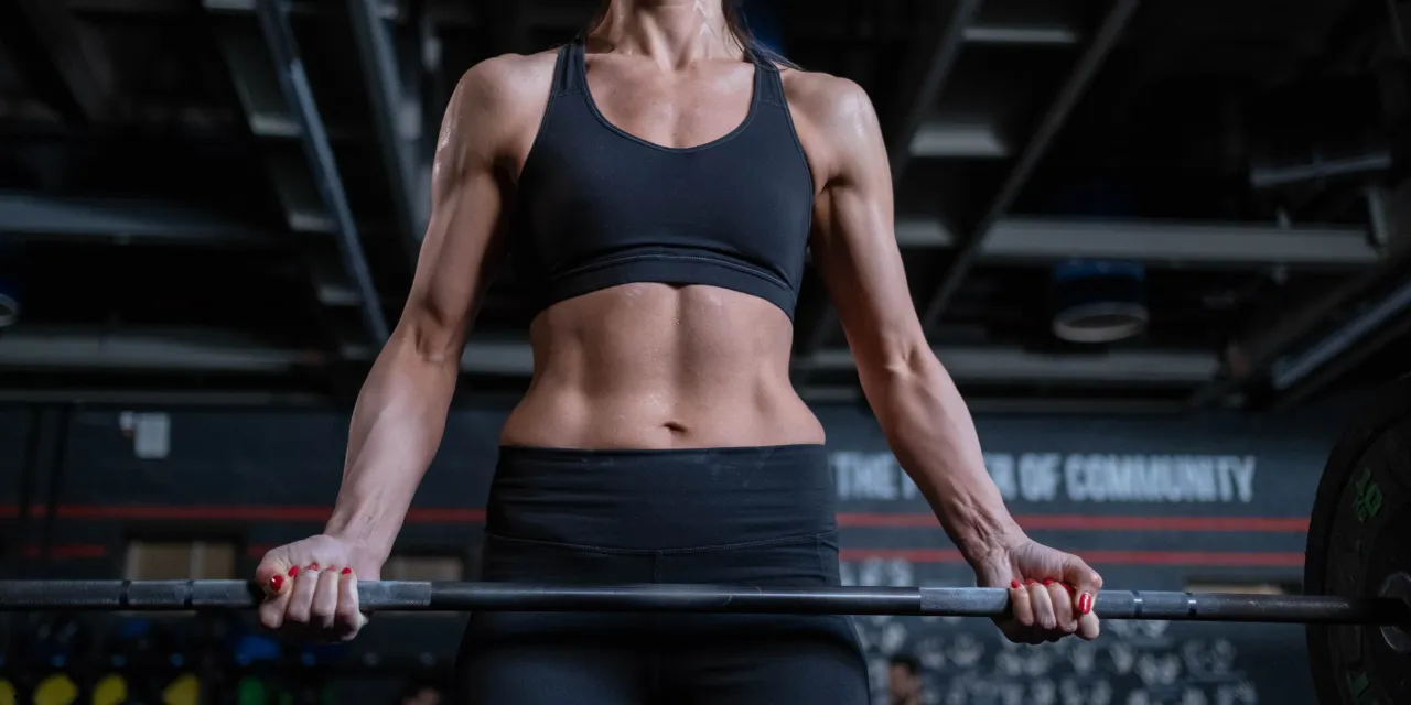 Kobieta z wyrzeźbionym brzuchem trzyma sztangę. Czy shake proteinowy przed czy po treningu pomoże jej w budowaniu mięśni?