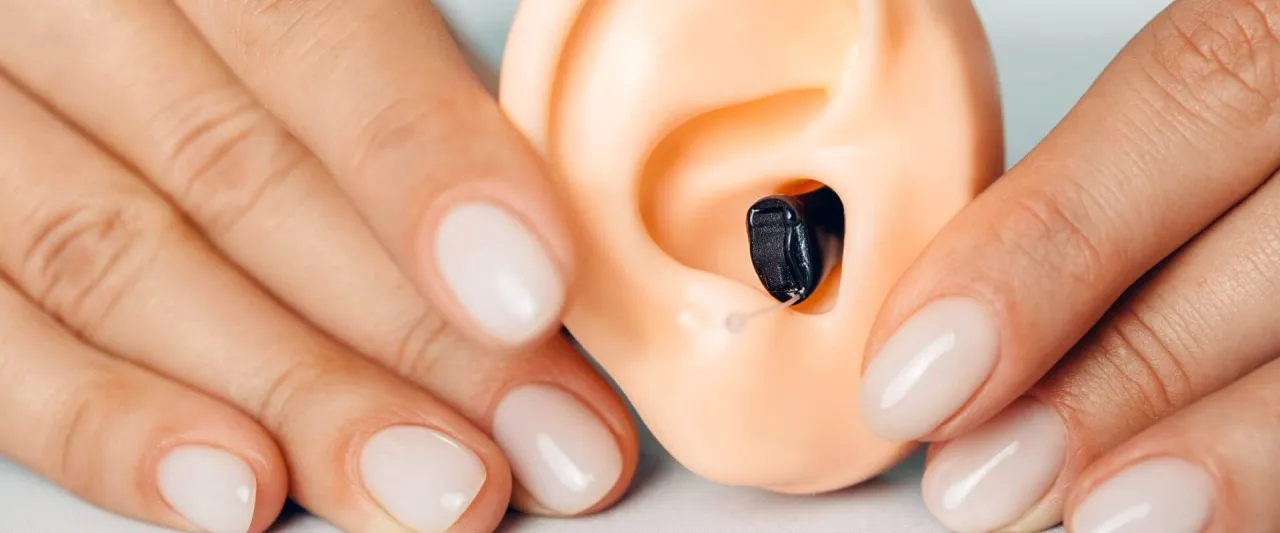 Dłonie zgrabnie umieszczają czarny aparat słuchowy w sztucznym uchu. Czasem szumi, gdy jest źle dopasowany.