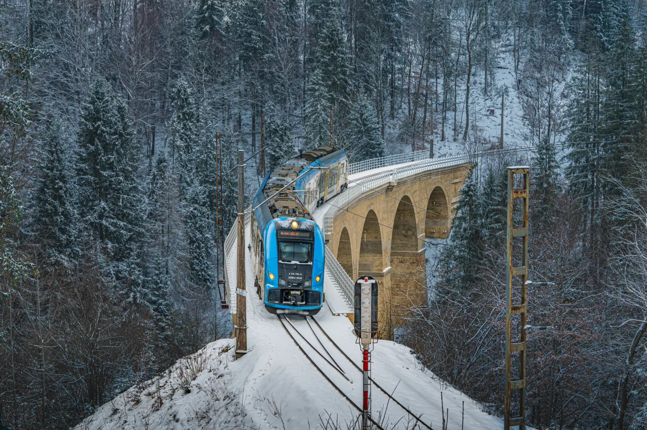 Niebieski pociąg mija kamienny wiadukt w zimowym lesie. Od kiedy pociągiem do Wisły, podr&oacute;ż przez ośnieżone drzewa jest magiczna.