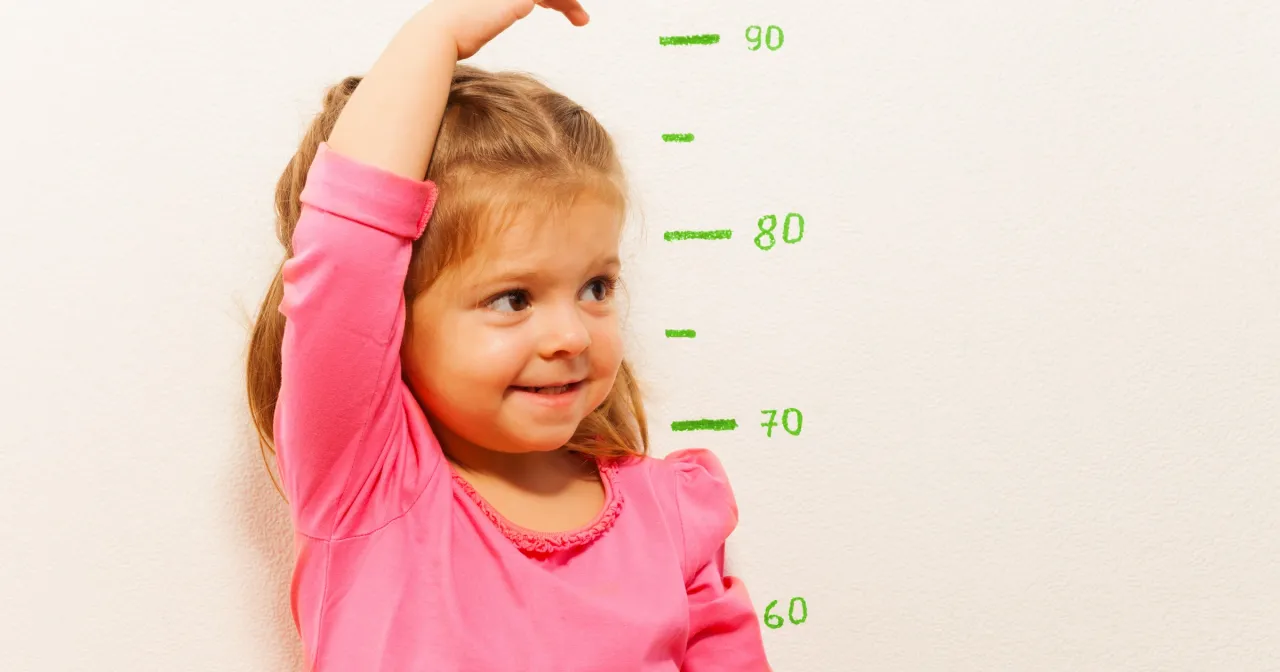 Mała dziewczynka w różowej bluzce mierzy swój wzrost przy ścianie z zaznaczonymi centymetrami. Zastanawia się, do którego roku życia się rośnie.
